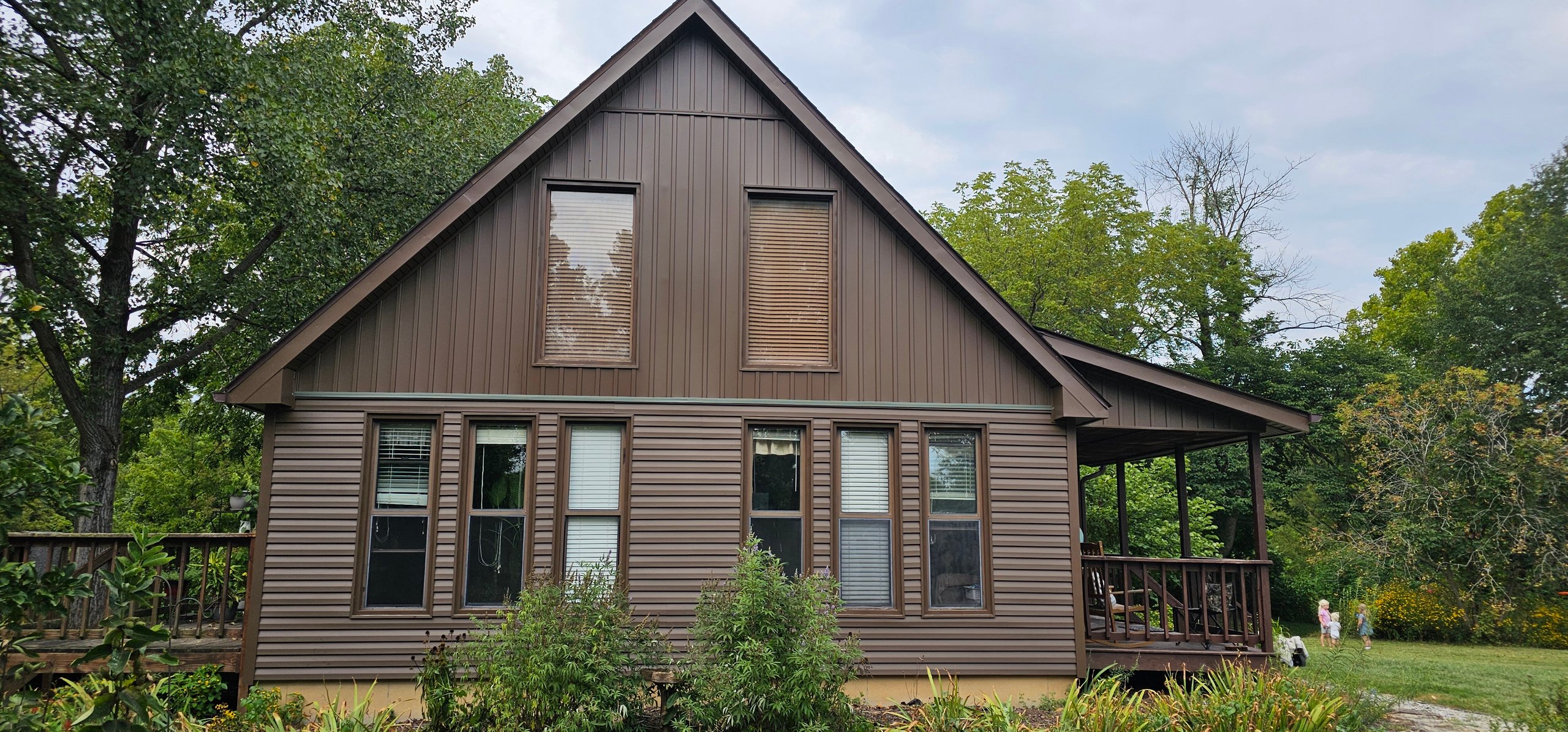 Brown wooden cabin with porch surrounded by green trees and garden